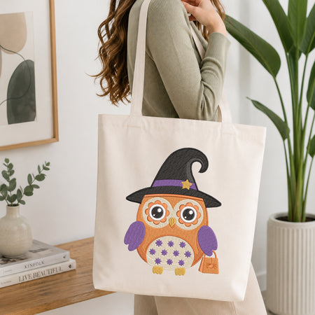 A woman wearing a sage green ribbed sweater carries a cream canvas tote bag with a colorful embroidered owl wearing a witch hat. The setting is a bright, modern room with a wooden table, books, a framed print, and a potted plant.