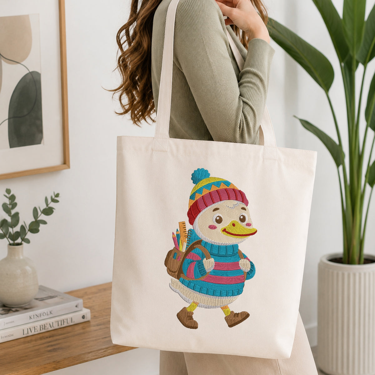 A woman wearing a sage green ribbed sweater carries a cream canvas tote bag with a colorful embroidered duck character wearing a striped beanie and blue sweater. The scene is set in a bright, modern room with a wooden table, books, a small plant, and a large green potted plant in the background.