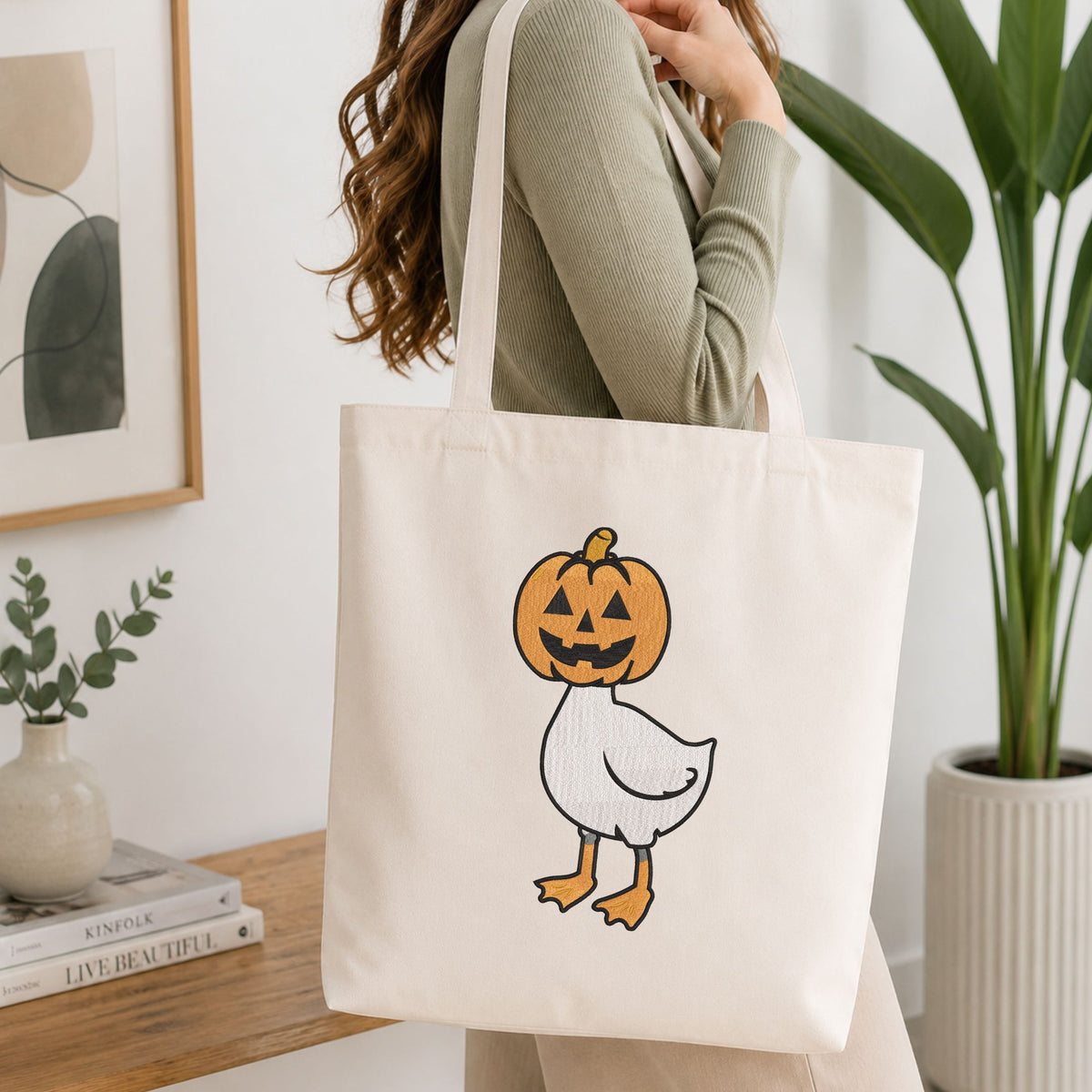 Woman carrying a white natural canvas tote bag featuring a cute duck in a pumpkin head Halloween machine embroidery design in a bright, modern interior setting
