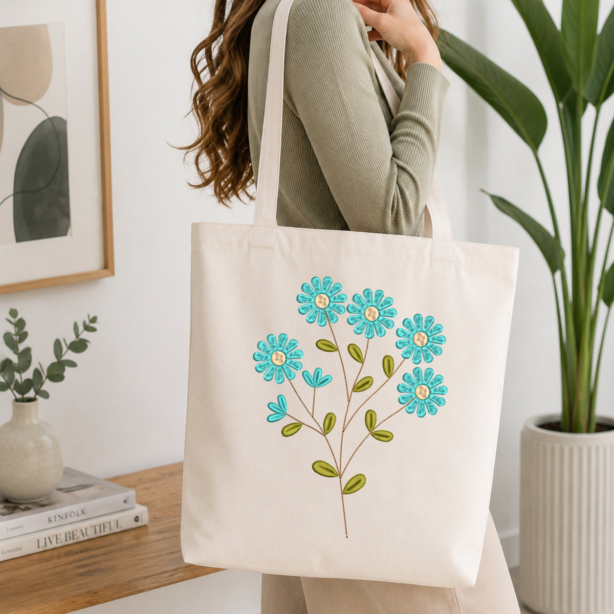 A woman carries a cream canvas tote bag with embroidered turquoise and green flowers. The photo is taken in a bright, modern room with a wooden table, books, a framed print, and a potted plant in the background.