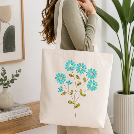 A woman carries a cream canvas tote bag with embroidered turquoise and green flowers. The photo is taken in a bright, modern room with a wooden table, books, a framed print, and a potted plant in the background.