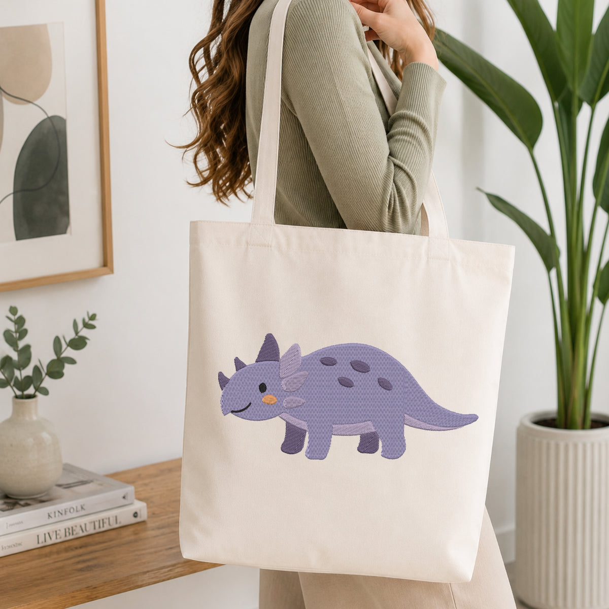 A woman wearing a sage green ribbed top carries a cream canvas tote bag with a purple cartoon Triceratops dinosaur printed on the front, set in a bright, modern room with a wooden table, books, and a potted plant.