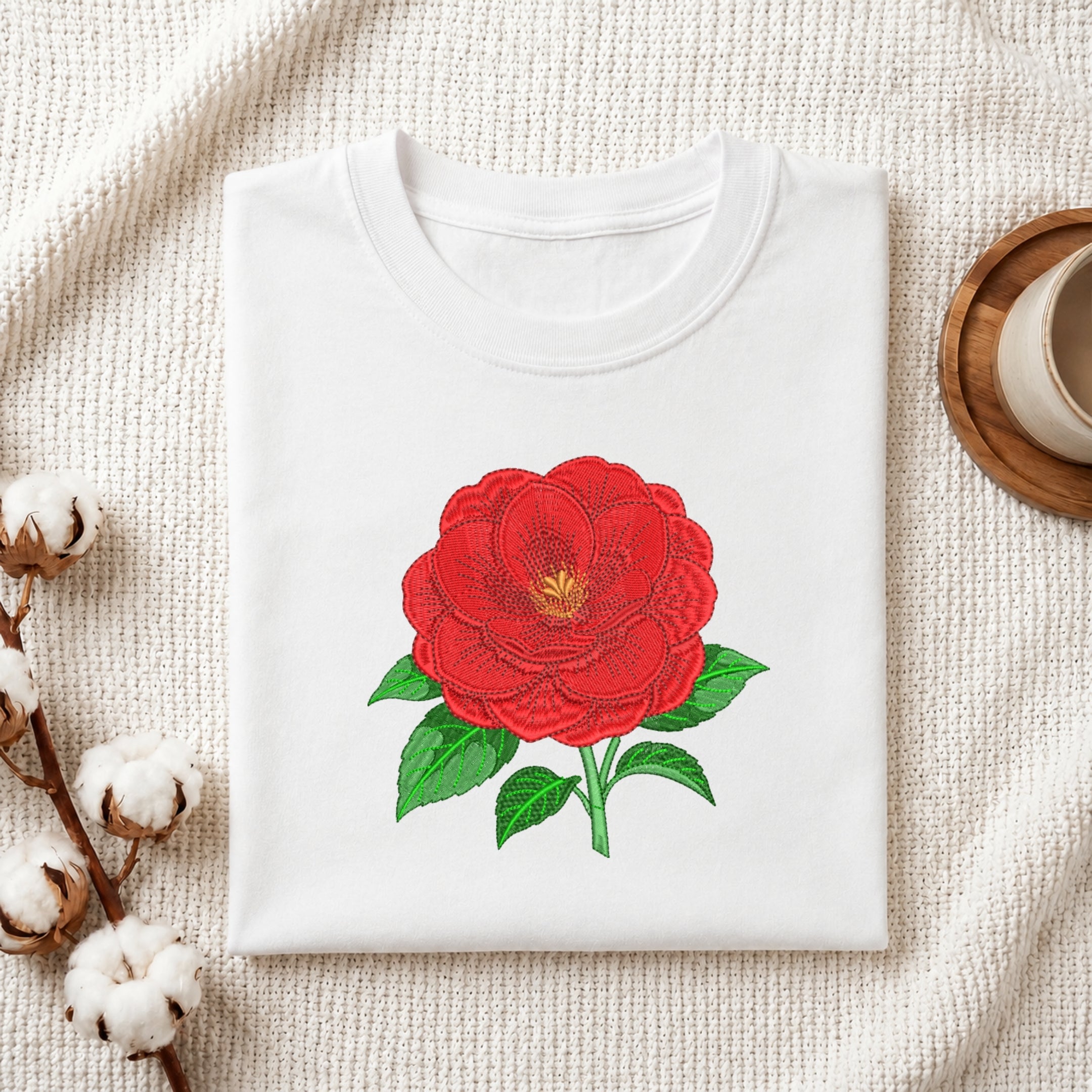 Folded white t-shirt mockup with a large red camellia floral embroidery design with green leaves.