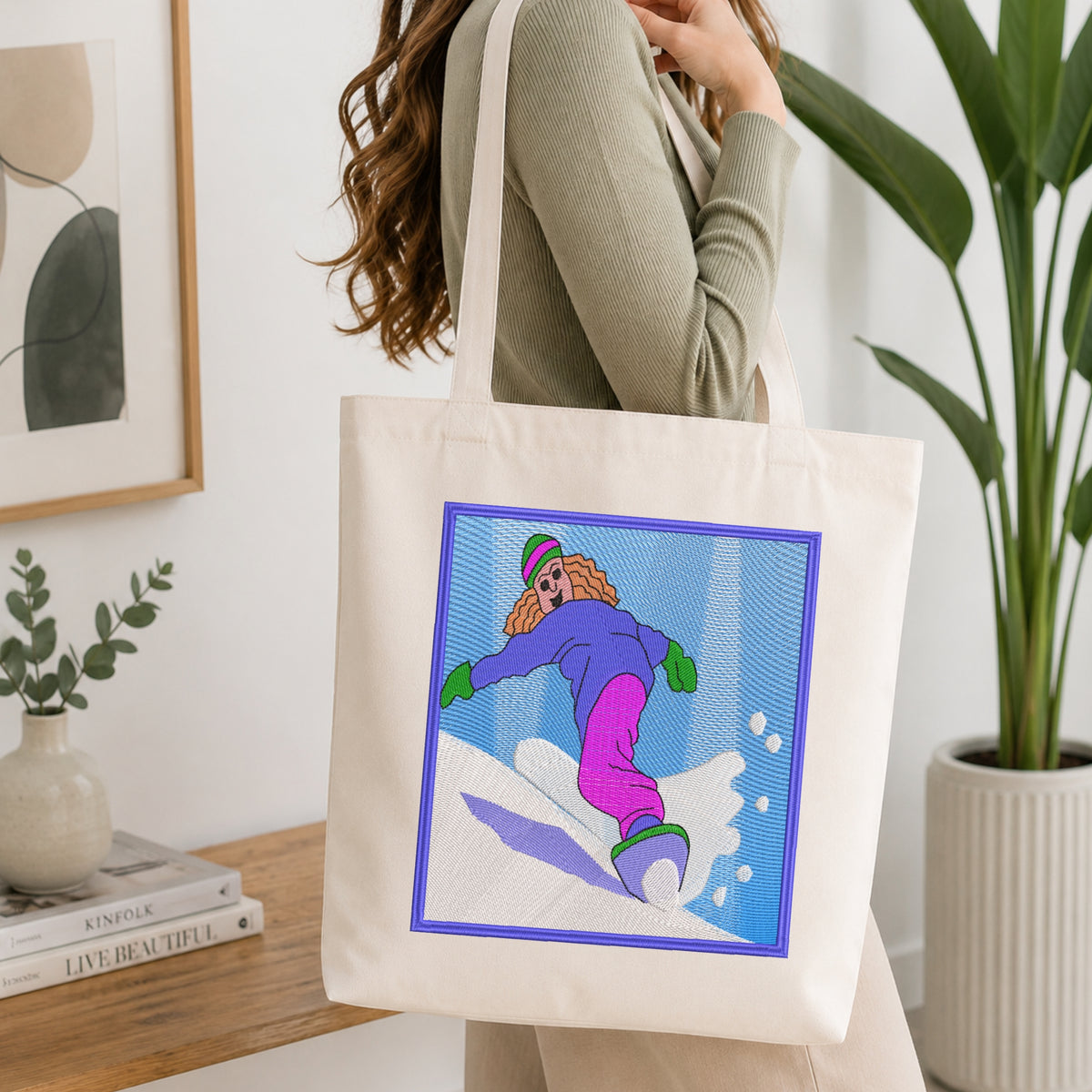 A woman wearing a sage green ribbed sweater carries a cream canvas tote bag with a colorful graphic print of a cartoon snowboarder. The setting is a bright, modern room with a wooden table, books, a potted plant, and framed wall art.