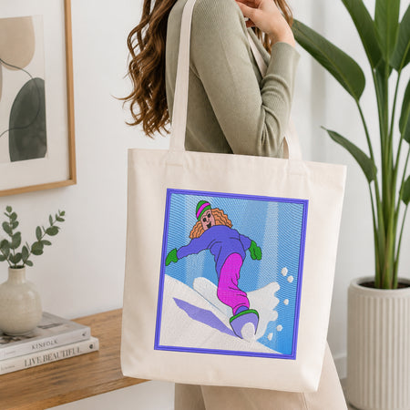 A woman wearing a sage green ribbed sweater carries a cream canvas tote bag with a colorful graphic print of a cartoon snowboarder. The setting is a bright, modern room with a wooden table, books, a potted plant, and framed wall art.