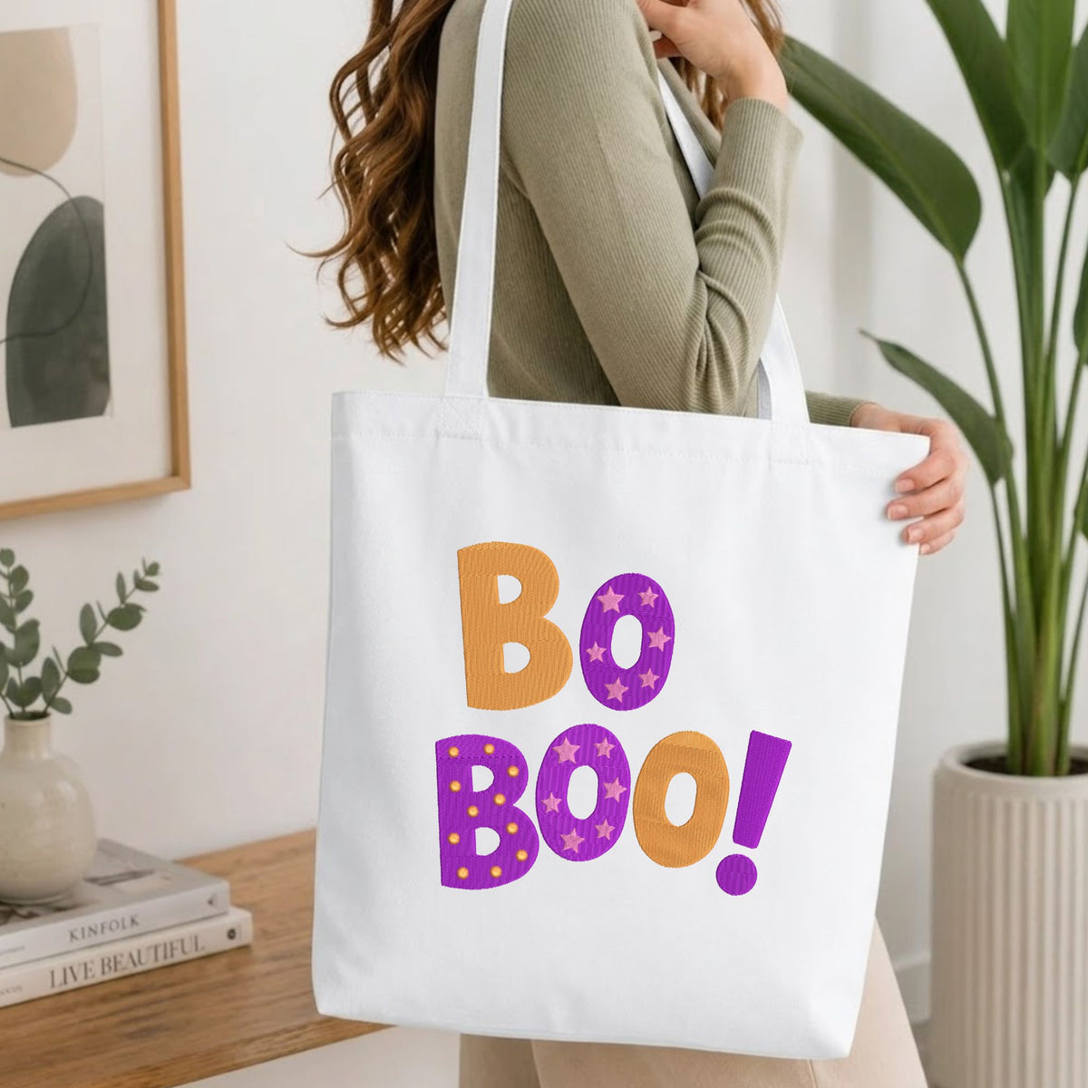 A woman wearing a sage green ribbed long-sleeve top carries a white canvas tote bag with the words 'Bo Boo!' printed in orange and purple. The photo is taken in a bright, modern room with a wooden table, books, a framed print, and a potted plant.