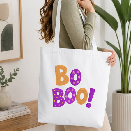 A woman wearing a sage green ribbed long-sleeve top carries a white canvas tote bag with the words 'Bo Boo!' printed in orange and purple. The photo is taken in a bright, modern room with a wooden table, books, a framed print, and a potted plant.