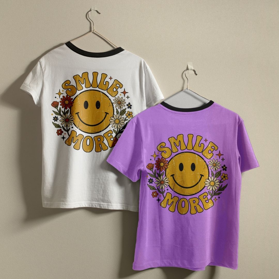 Two t-shirts, one white and one purple, hanging on a wall displaying a matching 70s style distressed smiley face and floral print.