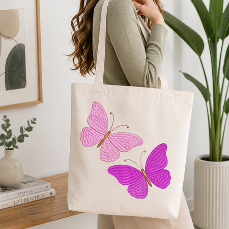 A woman wearing a sage green ribbed sweater carries a cream canvas tote bag with two large embroidered butterflies, one pink and one purple, against a bright indoor background with a plant and framed art.