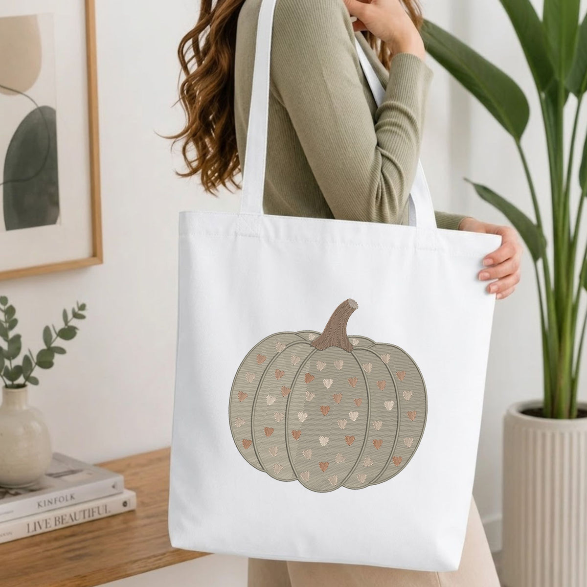 A woman wearing a sage green ribbed sweater carries a white canvas tote bag featuring a printed design of a pumpkin with hearts in beige, brown, and pink. The background is a bright, modern room with a wooden table, books, a plant, and framed wall art.