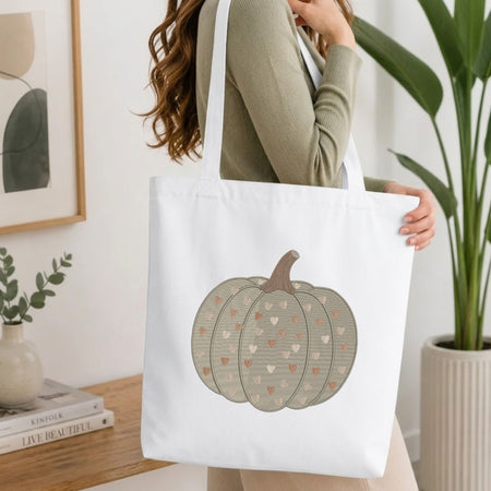 A woman wearing a sage green ribbed sweater carries a white canvas tote bag featuring a printed design of a pumpkin with hearts in beige, brown, and pink. The background is a bright, modern room with a wooden table, books, a plant, and framed wall art.