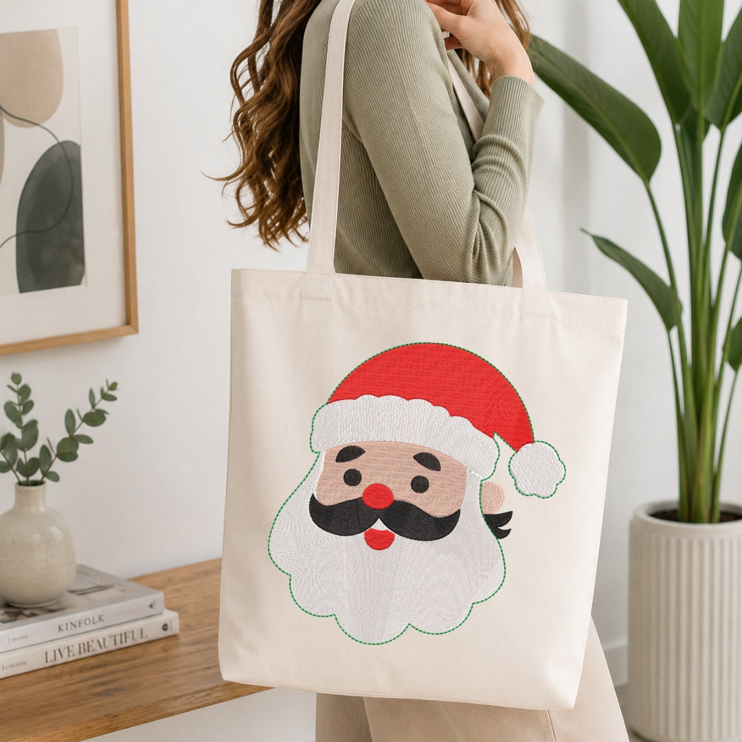 A woman in a ribbed sage green top carries a cream canvas tote bag featuring a large embroidered-style Santa Claus face with a red hat, white beard, black mustache, and red nose, set against a bright indoor background with a plant and books.