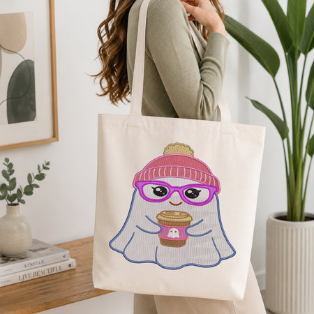 A woman wearing a sage green ribbed sweater carries a cream canvas tote bag. The bag features a cute cartoon ghost graphic wearing pink striped glasses, a pink beanie, and a white striped body, holding a coffee cup.