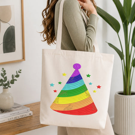 A person wearing a ribbed sage green top carries a cream canvas tote bag with a colorful rainbow party hat design and small stars, shown in a bright, modern room with a wooden table, books, and a potted plant.