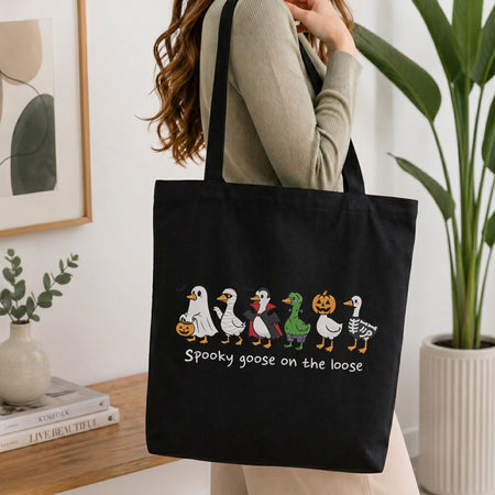  Woman carrying a black canvas tote bag featuring the "Spooky goose on the loose" Halloween costume geese machine embroidery design in a bright interior setting   