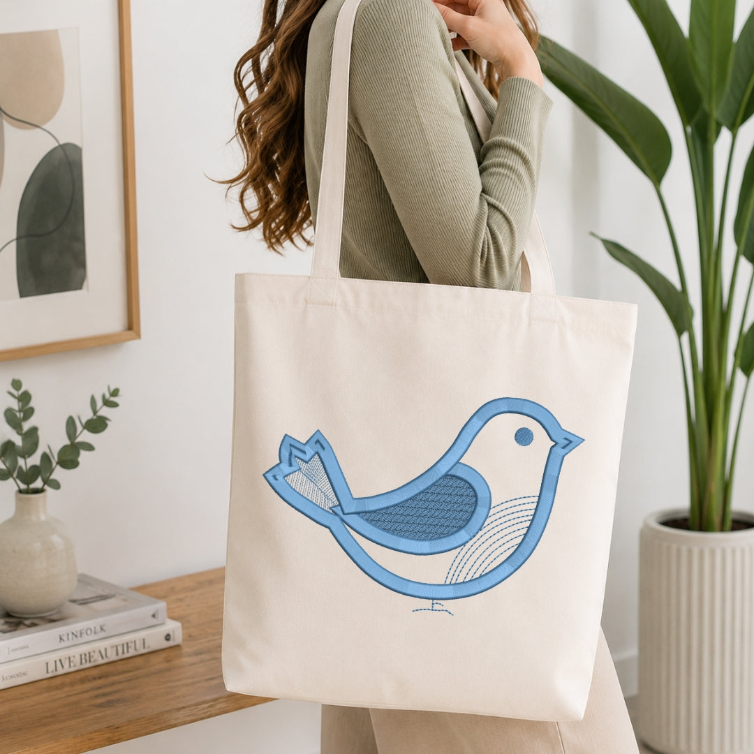 An off-white canvas tote bag with a blue bird graphic, carried on the shoulder of a woman wearing a sage green ribbed sweater in a bright, modern room with a wooden table and potted plants.