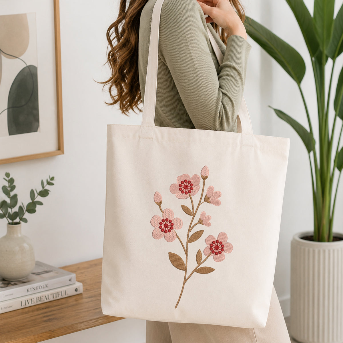A woman wearing a sage green ribbed sweater carries a cream canvas tote bag with a pink and brown floral embroidery design, set in a bright, modern room with a wooden table and potted plants.