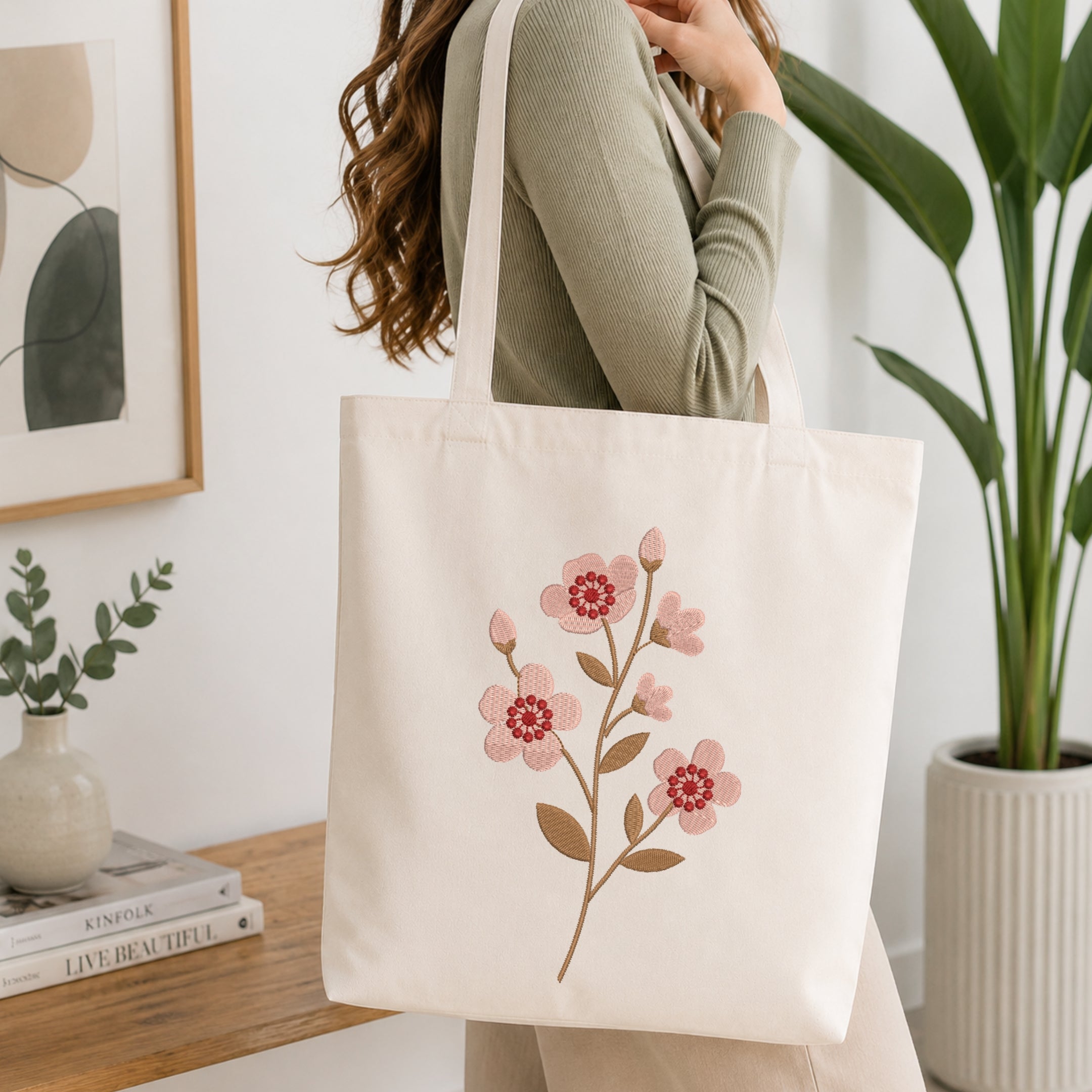 A woman wearing a sage green ribbed sweater carries a cream canvas tote bag with a pink and brown floral embroidery design, set in a bright, modern room with a wooden table and potted plants.