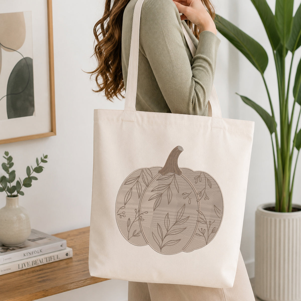 A woman wearing a sage green ribbed sweater carries a cream canvas tote bag with a large gray pumpkin illustration featuring leafy vine patterns. The scene is set in a bright, modern room with a wooden table, books, a small plant, and a potted green plant in the background.