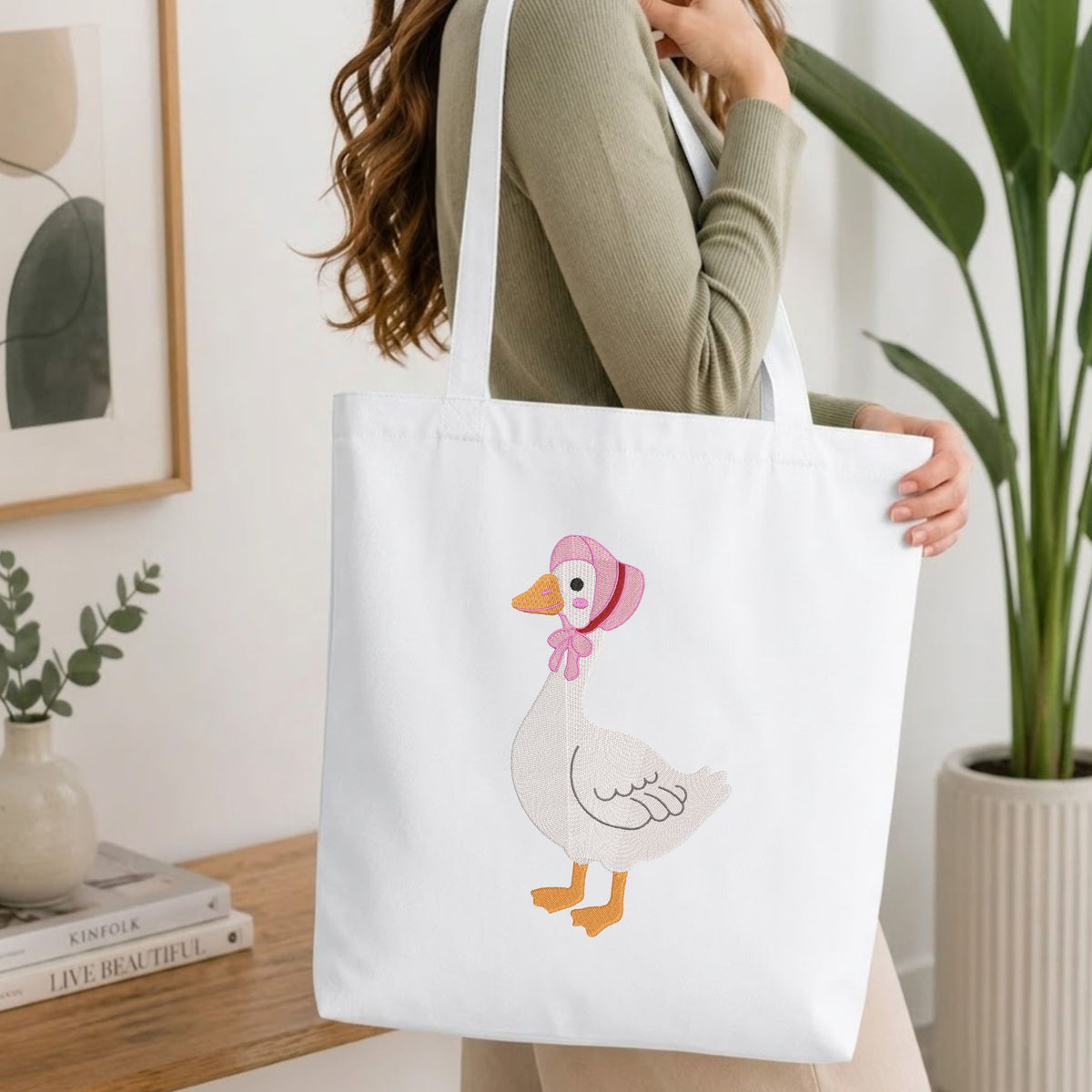 Woman carrying a white natural canvas tote bag featuring an embroidered cute goose in a bonnet design in a bright, modern interior setting.