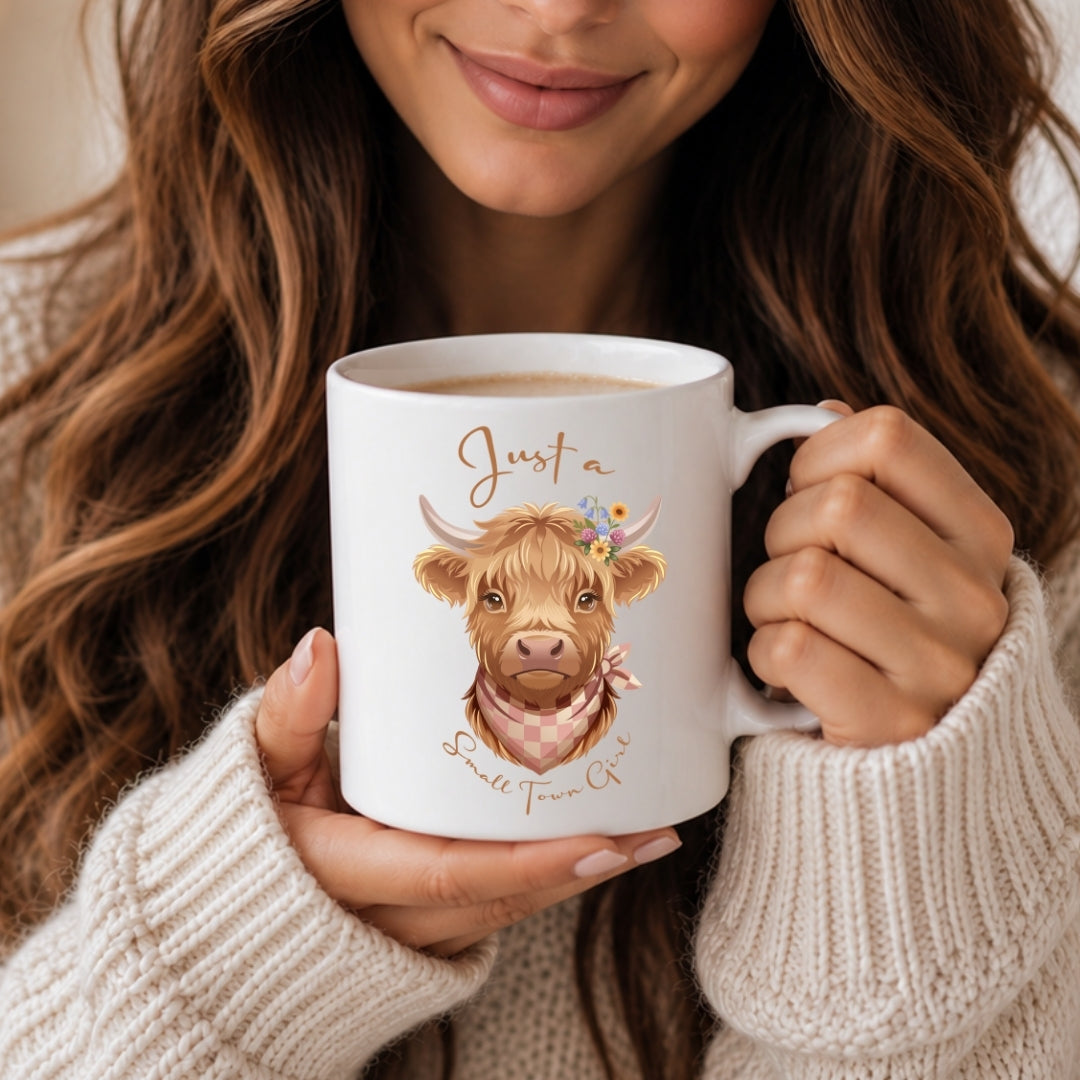A person in a cream knit sweater holds a white ceramic mug featuring a cute illustrated Highland cow with the text 'Just a Small Town Co.' The person is cropped from the mouth down, with long wavy brown hair visible around the mug.