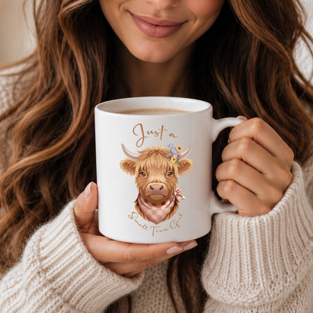 A person in a cream knit sweater holds a white ceramic mug featuring a cute illustrated Highland cow with the text 'Just a Small Town Co.' The person is cropped from the mouth down, with long wavy brown hair visible around the mug.