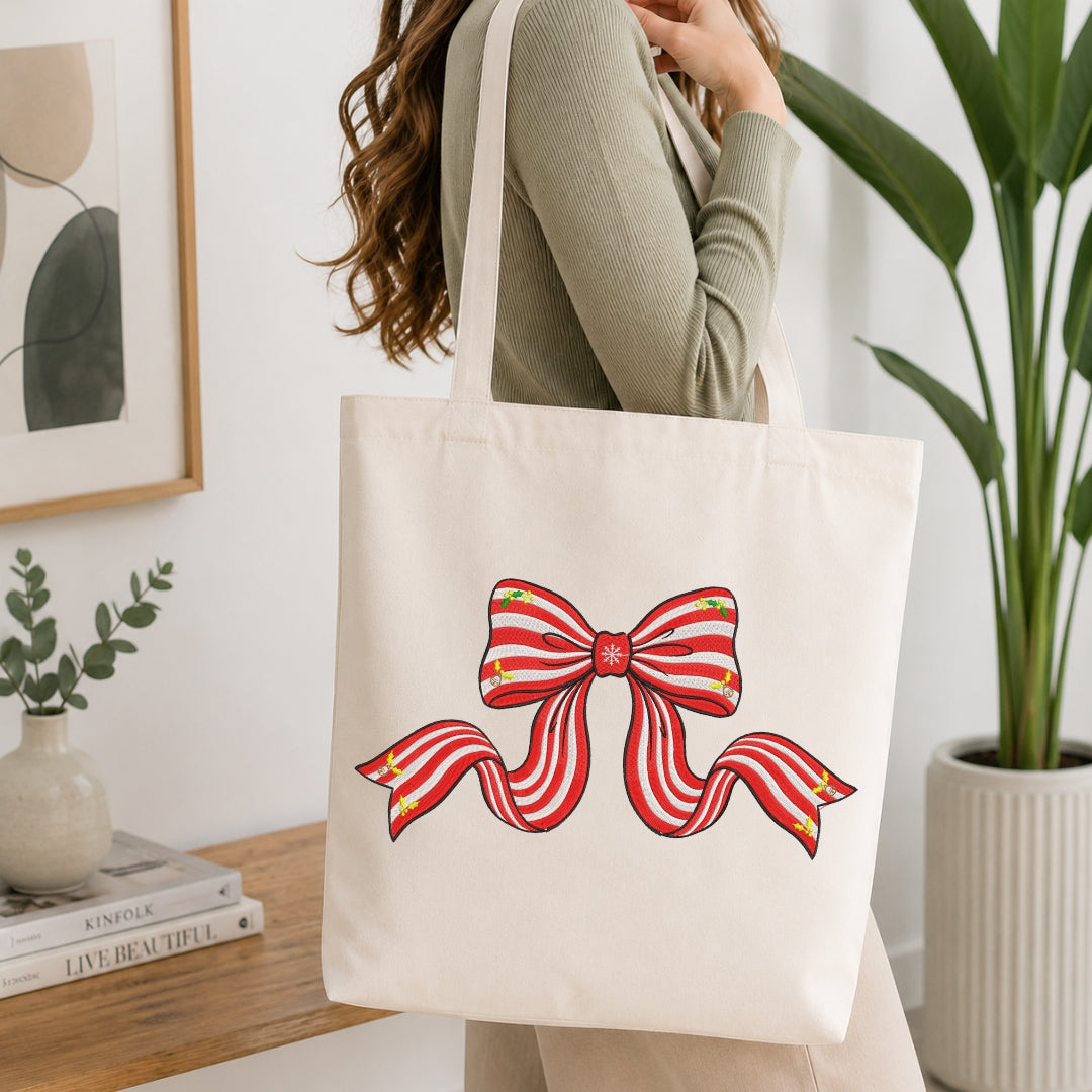 A person carrying a cream-colored tote bag featuring an embroidered design of a red and white striped holiday ribbon bow with small holly accents.