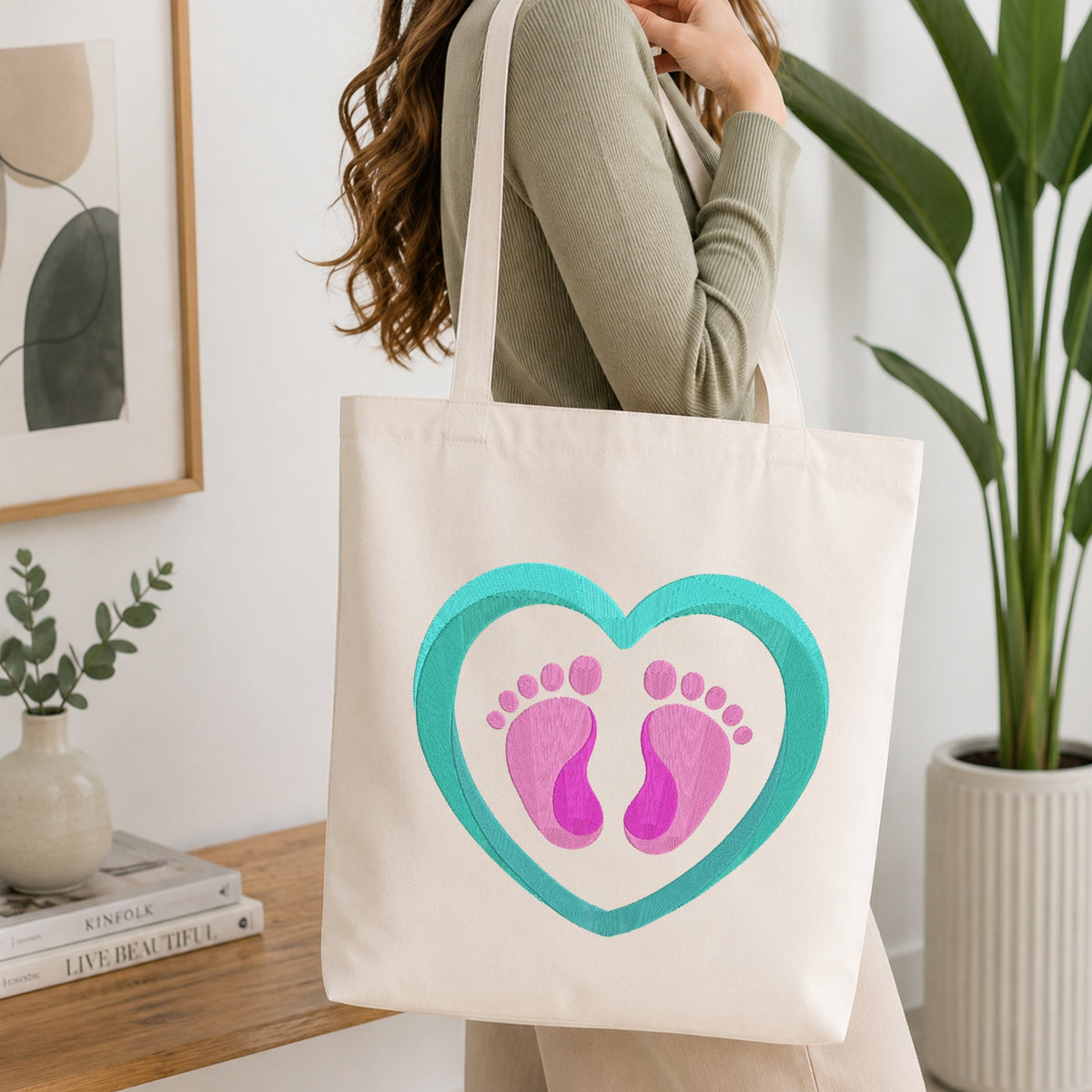 Woman carrying a white natural canvas tote bag featuring an embroidered baby feet in a heart design in a bright, modern interior setting.