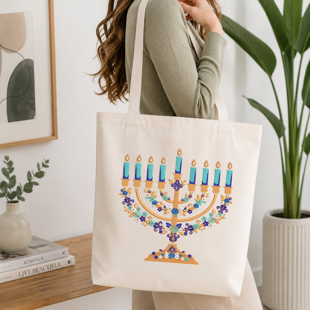 A woman wearing a sage green ribbed sweater carries a cream canvas tote bag featuring a colorful printed menorah design with blue candles and floral garlands, set in a bright, modern room with a wooden table and potted plants.