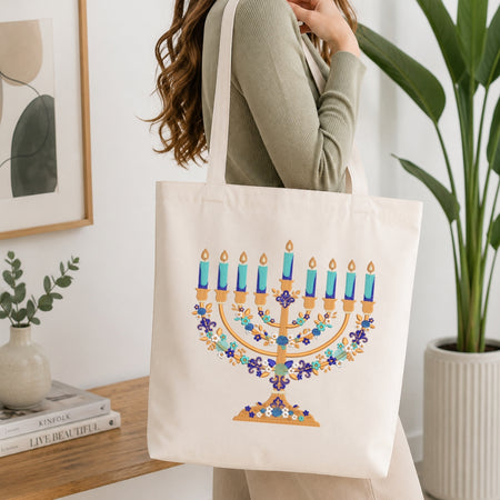 A woman wearing a sage green ribbed sweater carries a cream canvas tote bag featuring a colorful printed menorah design with blue candles and floral garlands, set in a bright, modern room with a wooden table and potted plants.