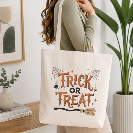 Woman carrying a white natural canvas tote bag featuring a "Trick or Treat" witch broom machine embroidery design in a bright, modern interior setting   