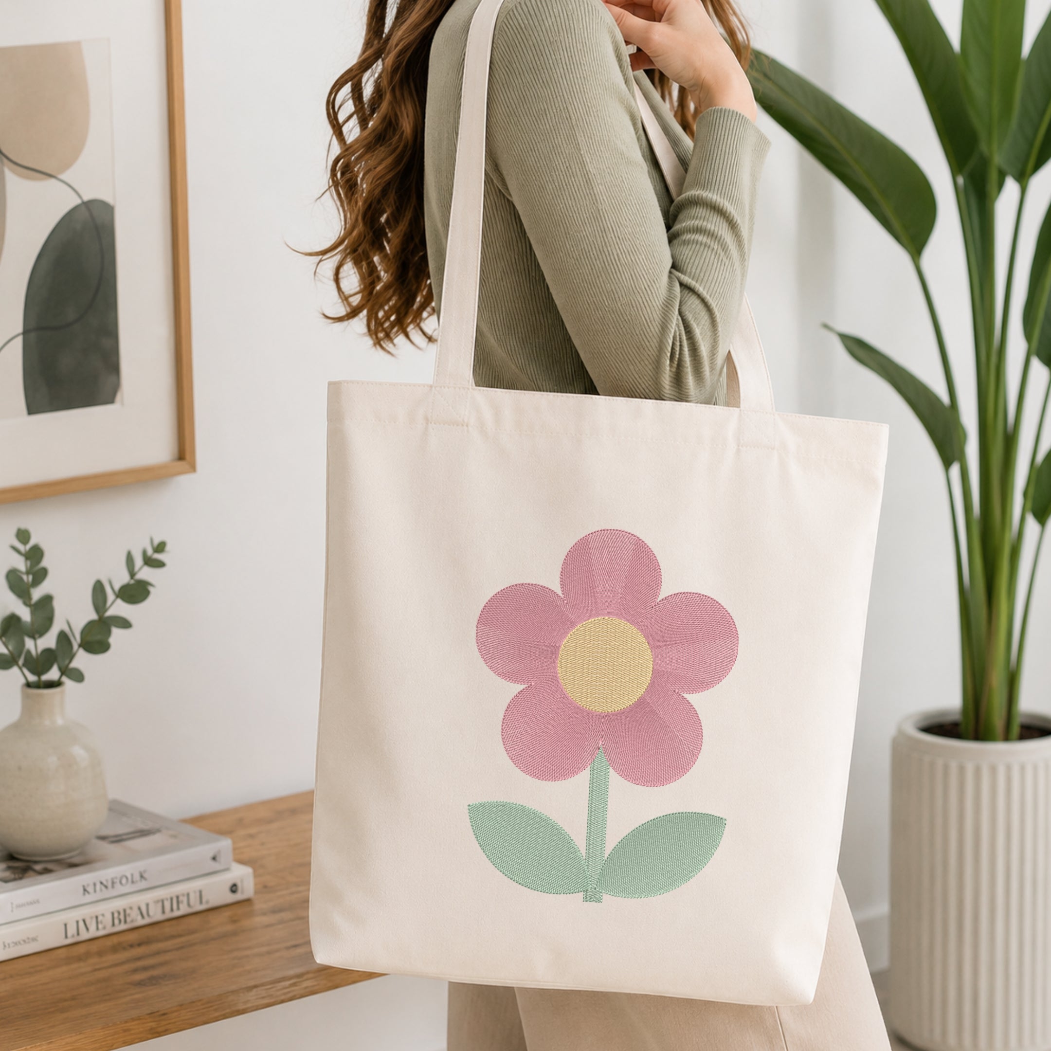 A woman wearing a sage green ribbed long-sleeve top carries a cream canvas tote bag with a large pink flower and mint green leaves printed on the front, set in a bright, modern room with a wooden table and potted plants.