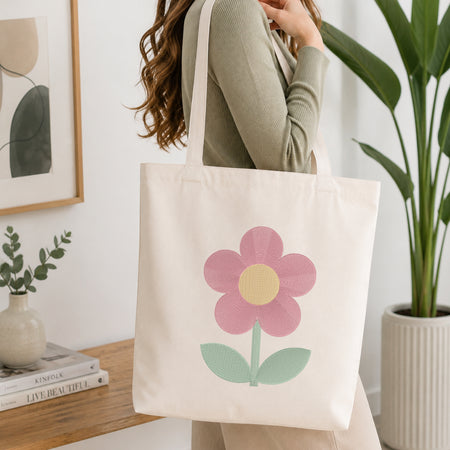 A woman wearing a sage green ribbed long-sleeve top carries a cream canvas tote bag with a large pink flower and mint green leaves printed on the front, set in a bright, modern room with a wooden table and potted plants.