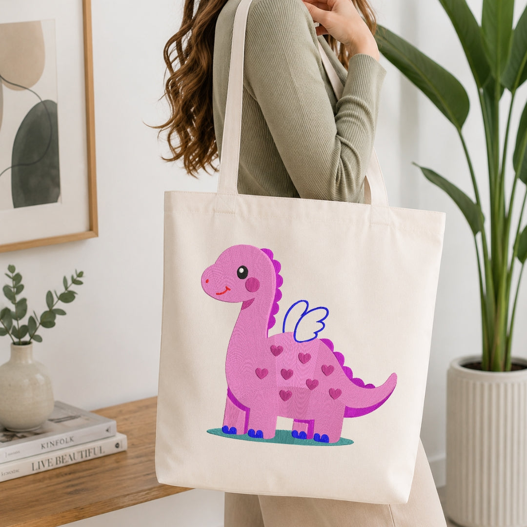 A woman wearing a sage green ribbed sweater carries a cream canvas tote bag with a cute pink winged dinosaur printed on the front, set in a bright, modern room with a wooden table, books, and a potted plant.