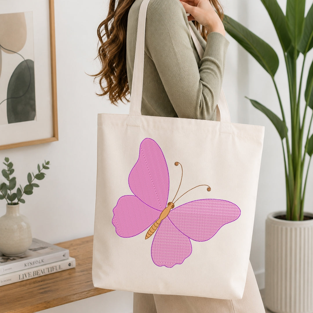 A woman wearing a sage green ribbed sweater carries a cream canvas tote bag with a large pink and orange butterfly graphic on the front, set in a bright, modern room with a wooden table, books, and a potted plant.
