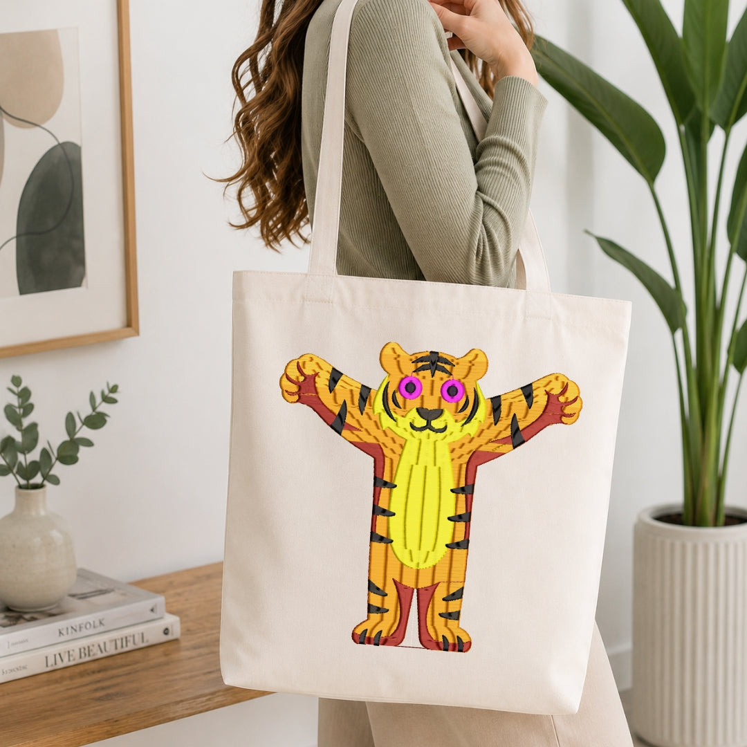 A woman wearing a sage green ribbed sweater carries a cream canvas tote bag featuring a colorful cartoon tiger illustration. The setting is a bright, modern room with a wooden table, books, a framed print, and a potted plant.