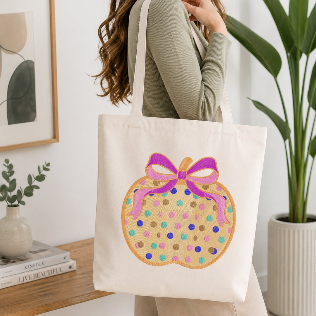 A woman wearing a sage green ribbed long-sleeve top carries a cream canvas tote bag. The bag features a graphic of a donut with colorful polka dots and a pink and purple bow.