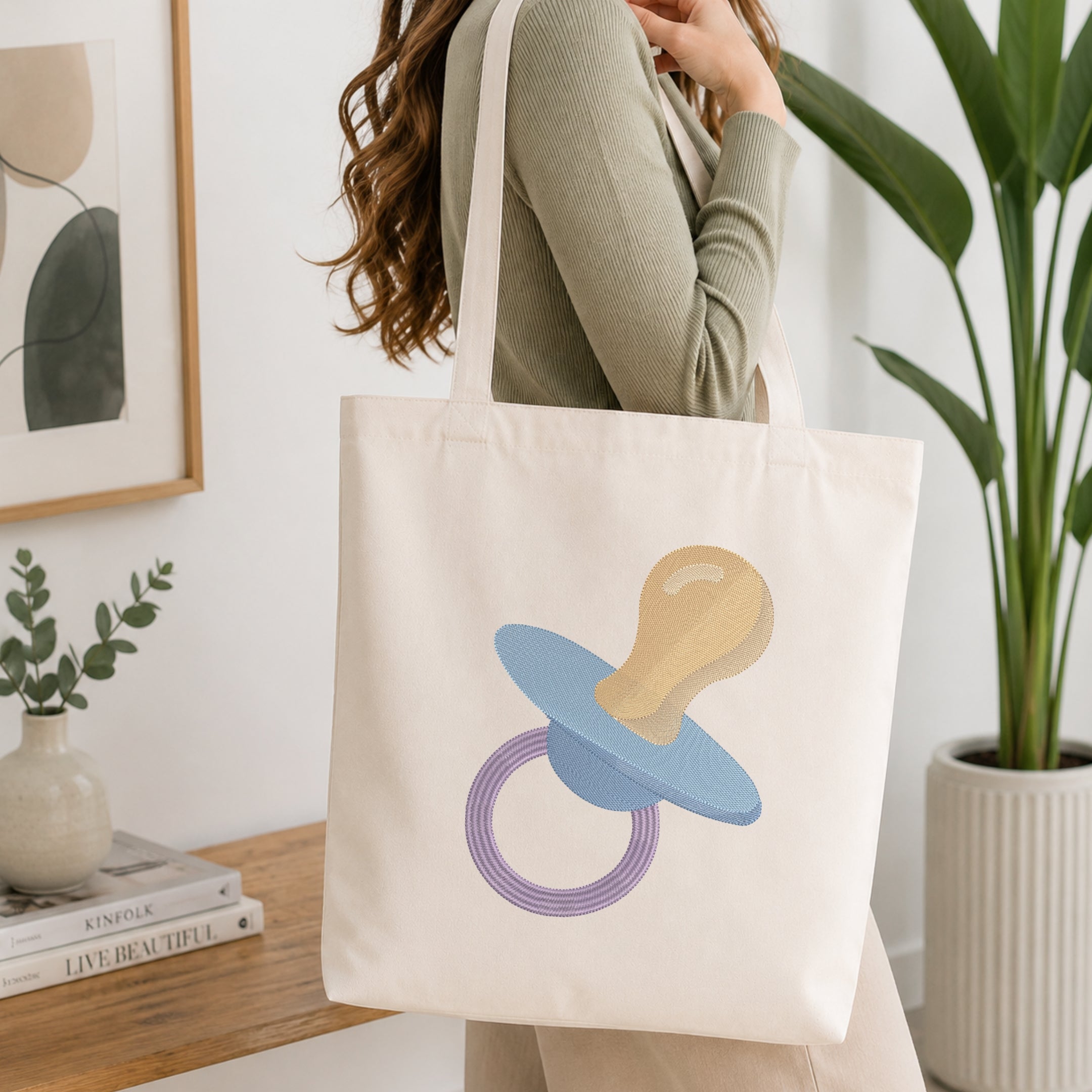 A woman wearing a sage green ribbed long-sleeve top carries a cream canvas tote bag with a pacifier graphic. The pacifier design features a light blue body, a beige nipple, and a purple ring.