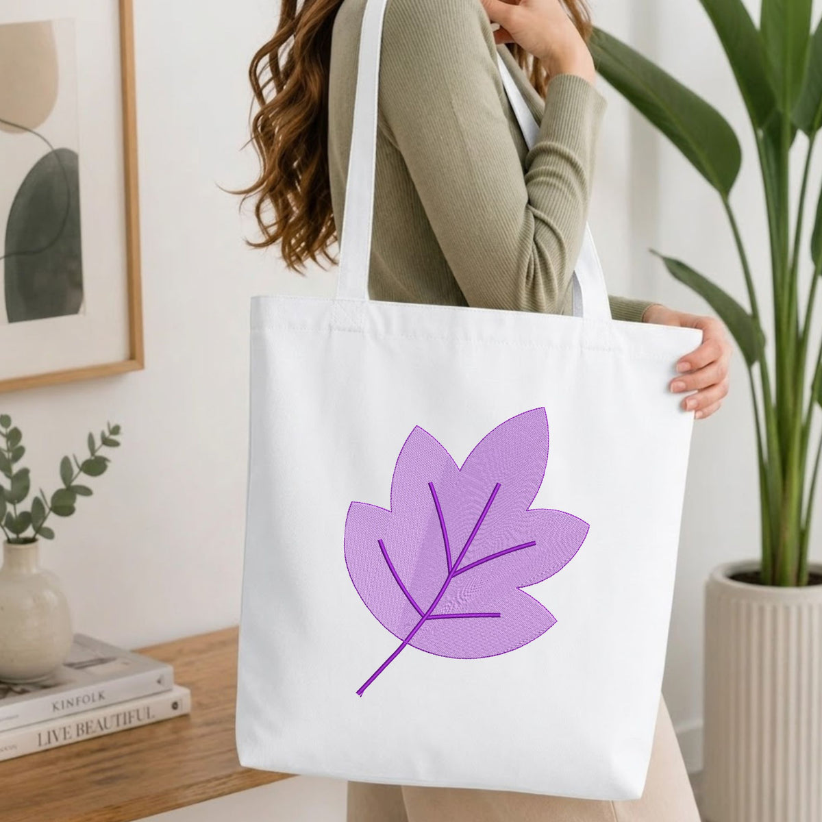 A woman wearing a sage green ribbed sweater carries a white canvas tote bag with a large purple leaf design printed on the front, set in a bright, modern room with a wooden table and potted plants.