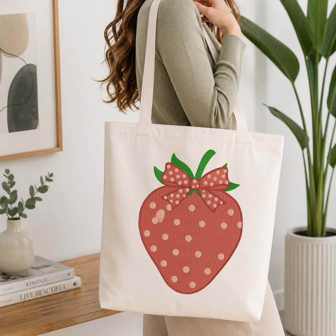 A woman wearing a sage green ribbed sweater carries a cream canvas tote bag with a large red strawberry graphic featuring white polka dots and a pink bow.