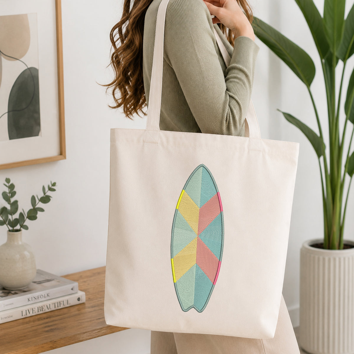 A woman wearing a sage green ribbed long-sleeve top carries a cream canvas tote bag with a colorful geometric surfboard design on the front, set in a bright, modern room with a wooden table, books, and a potted plant.