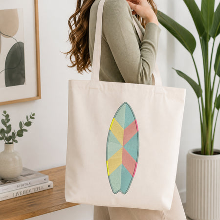 A woman wearing a sage green ribbed long-sleeve top carries a cream canvas tote bag with a colorful geometric surfboard design on the front, set in a bright, modern room with a wooden table, books, and a potted plant.