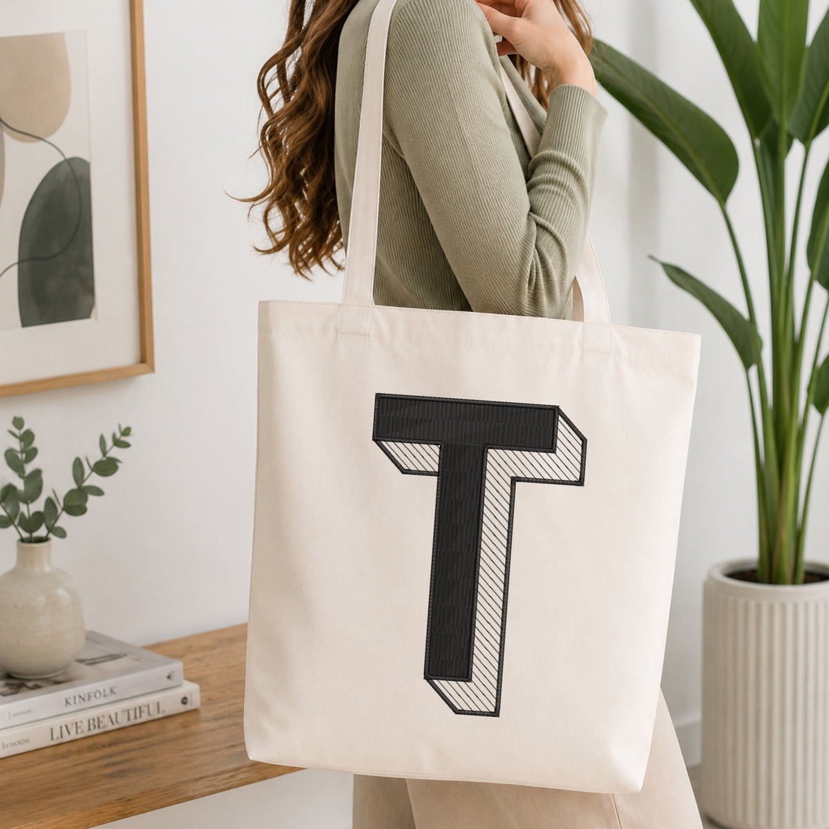 A woman wearing a sage green ribbed long-sleeve top carries a cream canvas tote bag with a large black embroidered letter 'T' on the front, set in a bright, modern room with a wooden table, framed art, and a potted plant.