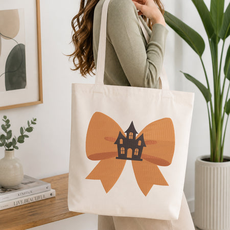 A person wearing a green long-sleeve top and carrying a natural canvas tote bag printed with the orange haunted house bow machine embroidery graphic.