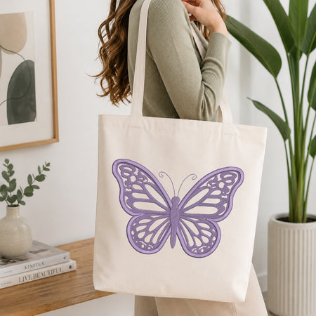 An off-white canvas tote bag with a purple embroidered butterfly design, carried by a woman wearing a sage green ribbed sweater in a bright, modern room with a wooden table and potted plants.