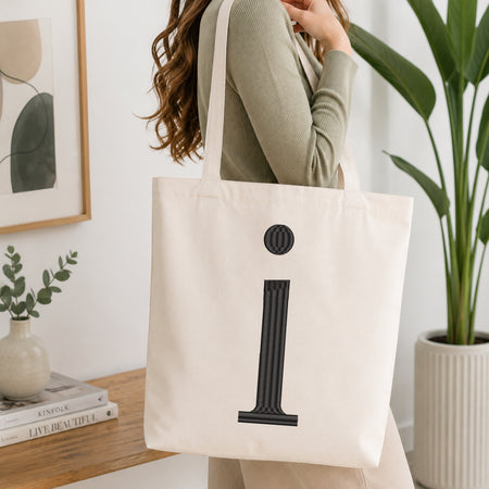 A woman wearing a sage green ribbed long-sleeve top carries a large off-white canvas tote bag with a bold black column-style letter 'i' graphic on the front, set in a bright, modern room with a wooden table, books, and a potted plant.