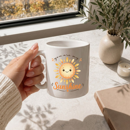 A person holding a white ceramic mug featuring a happy cartoon sun graphic and the text "you are my Sunshine".