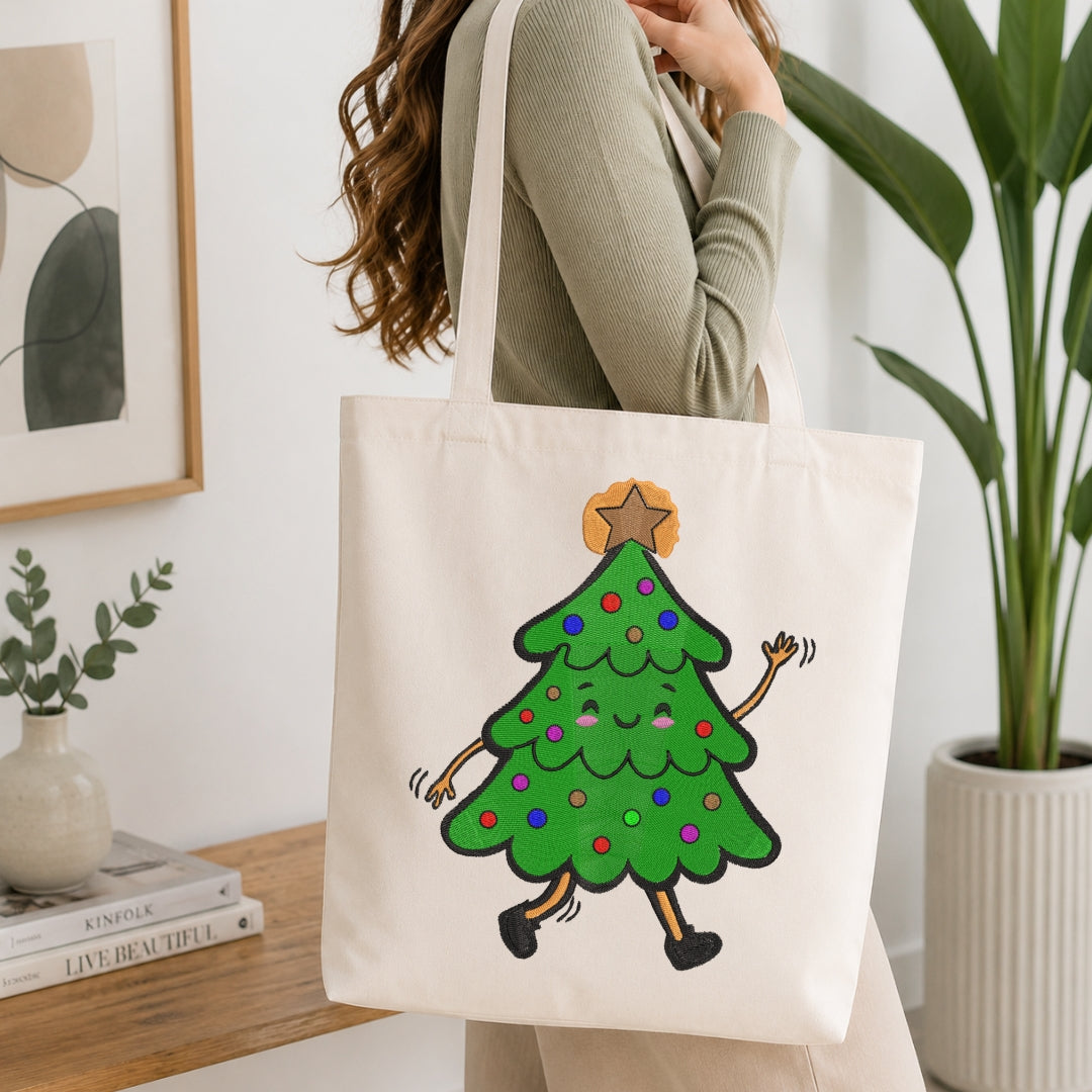 A woman wearing a sage green ribbed sweater carries a cream canvas tote bag with a cartoon green Christmas tree graphic on the front, set in a bright, modern room with a wooden table, books, and a potted plant.