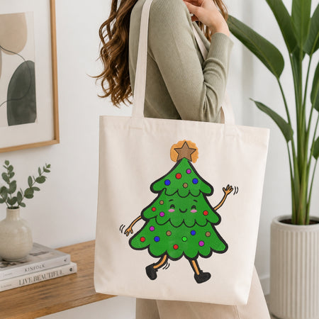 A woman wearing a sage green ribbed sweater carries a cream canvas tote bag with a cartoon green Christmas tree graphic on the front, set in a bright, modern room with a wooden table, books, and a potted plant.