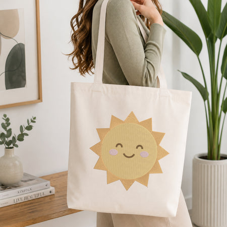 A woman wearing a sage green ribbed long-sleeve top carries a cream canvas tote bag with a large embroidered yellow sun face on the front, set in a bright, modern room with a wooden table, books, and a potted plant.